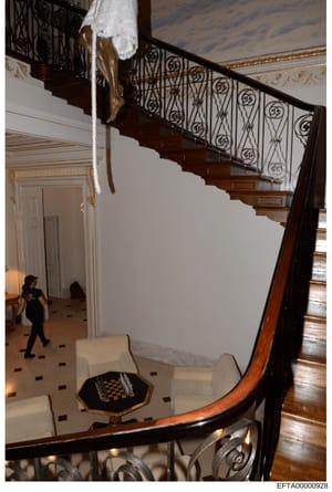 This evidence photograph shows the interior of a luxurious residence, depicting a two-story foyer with an ornate wooden staircase featuring decorative iron railings, marble floors with dark inlays, classical doorways, and high ceilings with crown mol