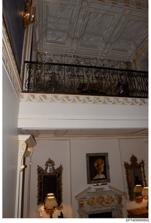 This is an interior photograph of a luxuriously decorated room featuring neoclassical architectural details, including an ornate coffered ceiling with gold leaf detailing, a wrought iron mezzanine railing with scrollwork patterns, decorative mirrors 