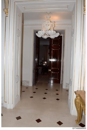 This is an evidence photograph showing an elegant hallway interior featuring a white and gold decorative scheme. The hallway displays a sophisticated chandelier with white petal-like elements, white-trimmed doorways, gold leaf accents, a marble floor