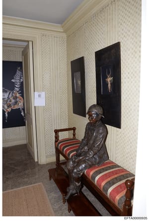 This is a photograph of an interior room, likely from one of Jeffrey Epstein's properties, showing a hallway or bedroom area with decorative elements. The room features cream-colored wallpapered walls, dark framed artwork depicting animals, a bronze 