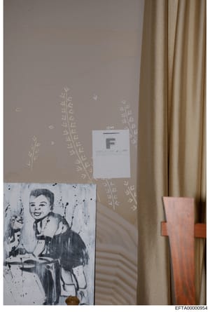 This is an evidence photograph showing an interior wall with decorative wallpaper featuring plant motifs, a tan curtain on the right, and two items: a white evidence marker labeled 'F' and what appears to be a black and white artwork or print of a sm