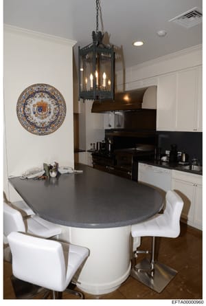 This photograph shows a modern kitchen interior featuring a curved island with gray countertop and white cabinetry, white modern bar seating, black appliances, ornate blue and gold decorative plate on the wall, and a black iron pendant light fixture.