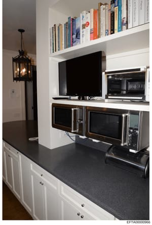 This is a photograph of a modern kitchen interior featuring built-in cabinetry, shelving, and entertainment equipment. The image shows a white and dark kitchen counter with multiple microwaves, a flat-screen television, and bookshelves displaying var