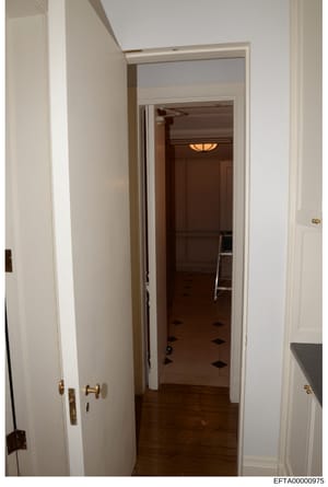 This is a photograph of an interior hallway in a residential property, showing white-painted walls, wooden flooring, multiple doorways with brass hardware, and interior lighting. The image appears to be part of evidence documentation for the investig