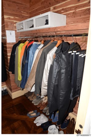 This is a photograph of a residential closet interior showing clothing items hanging on a metal rod and various shoes and accessories stored on the floor. The closet features wood paneling walls and ceiling, white cabinet storage above, and a dark wo