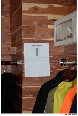 This is a forensic evidence photograph showing the interior of a wood-paneled room with a closet, clothing on hangers, and an evidence collection marker placed on the wall. The marker appears to be used to document and scale evidence during an invest