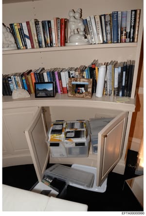 This photograph shows a built-in bookcase and storage unit containing approximately 100+ books, framed photographs, decorative objects, and storage containers. The image appears to be evidence documentation from a property search, showing the interio