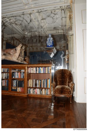 This photograph documents an elegantly appointed library or study room featuring high ceilings with ornate architectural detailing, built-in wooden bookshelves, fine art objects, and luxury furnishings. The room exhibits classical interior design wit
