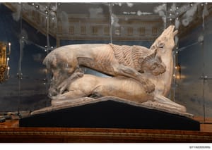This photograph documents a classical or neoclassical stone sculpture depicting a reclining mythological creature (possibly a dragon or chimera) displayed in a museum-quality glass case. The artifact is mounted on a dark base within a climate-control