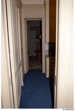 This is a photograph of an interior hallway featuring white paneled doors on both sides, blue carpeting, and white trim. The hallway appears to lead to additional rooms with darker interior spaces visible through open doorways. The image documents th