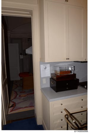 This is an evidence photograph showing the interior of a residential room, featuring a white dresser with gray countertop displaying multiple wooden jewelry boxes with glass covers. A doorway to an adjoining room is visible on the left. The photo app