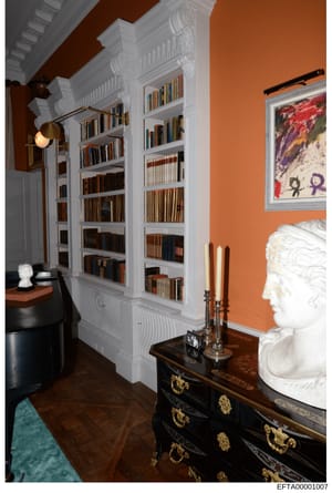 This is a photograph of an elegantly appointed library or study room, featuring floor-to-ceiling white built-in bookshelves filled with books, ornate classical architectural details, and high-end furnishings. The room displays orange/rust-colored wal
