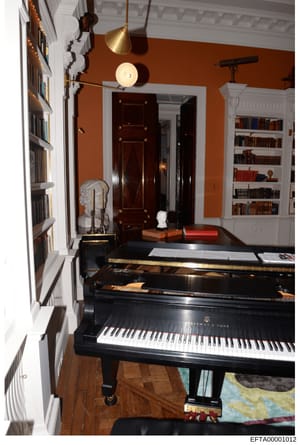 This is a photograph of an elegantly decorated music room featuring a black Steinway & Sons grand piano as the centerpiece. The room displays sophisticated interior design with orange-painted walls, white crown molding, extensive built-in bookshelves