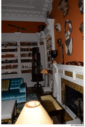 This is a photograph of an elegant interior room featuring built-in white bookshelves, a fireplace, and an extensive collection of African masks and art mounted on burnt-orange walls. The room displays high-end furnishings including a teal velvet cha