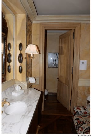 This is a photograph of an upscale bathroom interior, likely from one of Jeffrey Epstein's properties. The image shows high-end finishes including marble countertops, gold-plated fixtures, ornate wall sconces, and custom cabinetry. The bathroom is la