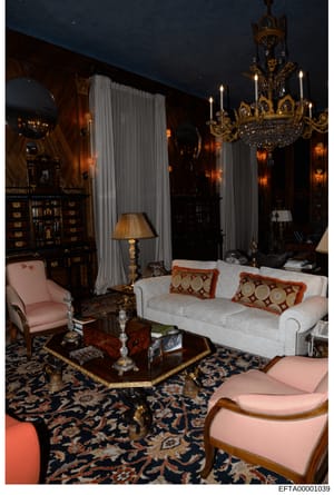 This photograph depicts an ornately decorated bedroom featuring high-end furnishings, including a white upholstered sofa with decorative pillows, pink wingback chairs, a brass chandelier, heavy drapery, antique wooden cabinetry, and elaborate gold-le