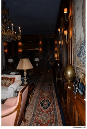 This is a photograph of an elegant interior hallway or gallery featuring high-end furnishings and décor. The space displays ornate chandeliers, wall sconces, decorative furniture including upholstered chairs, fine rugs, and carefully curated lighting