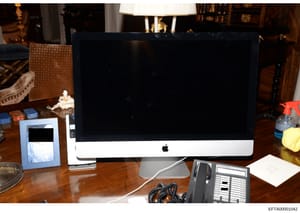 This is an evidence photograph showing a desk workspace with an Apple iMac computer and various office equipment and personal items. The image was taken as part of the investigation documentation, likely from one of Epstein's properties. The photogra