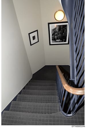 This is a photograph of an interior stairwell in what appears to be a residential or commercial property. The image shows a narrow staircase with dark gray carpeting, white walls, framed black-and-white artwork, a round wall sconce, and blue curtains