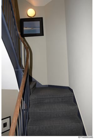 This is a photograph of an interior stairwell, likely from one of Epstein's properties. The image shows architectural details including a wooden handrail, dark carpeted stairs, white walls, recessed lighting, and a framed artwork depicting a sailboat