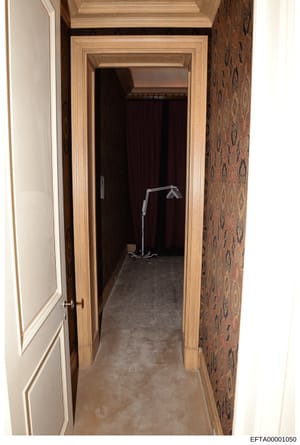 This is a photograph of an interior hallway featuring ornate wood-trimmed doorways, patterned wallpaper on the right wall, and concrete flooring. The image appears to be evidence documentation from a property associated with the investigation, showin