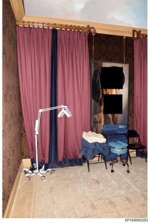 This photograph documents an interior room space featuring floor-to-ceiling pink and navy curtains on a brass rod, ornate patterned wallpaper, and what appears to be medical or examination equipment including a mobile light fixture and chairs with fo