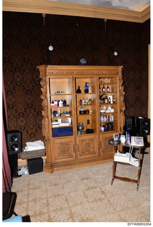 This photograph depicts an interior room featuring a large ornate wooden shelving unit displaying various luxury items, cosmetics, and decorative objects. The room contains premium audio equipment, damask-patterned wallpaper, and upscale furnishings.