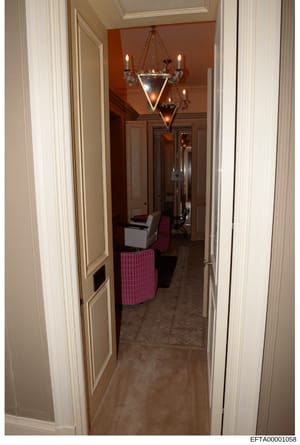 This is a photograph of an interior hallway in what appears to be a luxury residence. The hallway features rust/terracotta-colored walls, cream-colored trim, ornate lighting fixtures including a distinctive geometric chandelier, and upholstered furni