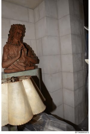 This is a photograph of an interior space showing a terracotta or brown-colored decorative bust sculpture positioned on a pedestal or furniture piece with a cream-colored fabric skirt. The background features white subway tile walls. The image appear