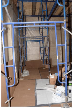 This is a photograph of an interior space under construction, showing blue metal framing structures, exposed wooden ceiling elements, cardboard boxes, and white materials scattered on the floor. The image appears to be documentation of ongoing renova