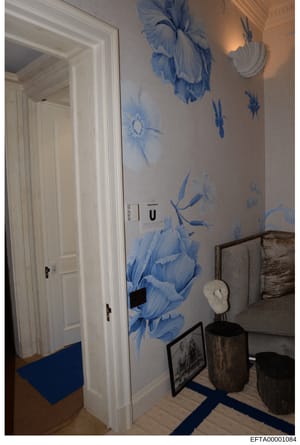 This is an evidence photograph of an interior room showing decorative wall art with blue floral motifs, white trim doors, and contemporary furnishings. The room appears to be part of a residence under investigation, with visible thermostat and framed