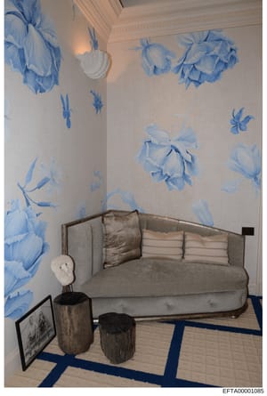 This photograph documents an interior room, likely from one of Epstein's properties, featuring distinctive blue and white botanical wallpaper with floral motifs, a gray upholstered sofa with decorative pillows, wooden accent pieces, and a geometric p