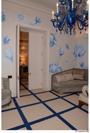 This is a photograph of an upscale interior space featuring decorative wallpaper with blue floral patterns, a blue crystal chandelier, cream-colored furniture, and a geometric floor pattern with blue accents. The room appears to be part of a high-end