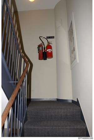 This is a photograph of an interior stairwell in a multi-story building, showing the staircase landing between floors with fire safety equipment mounted on the wall. The image captures architectural details and building safety infrastructure, likely 