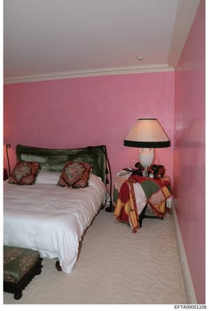 This is a photograph of a residential bedroom featuring pink-painted walls, a metal-framed bed with decorative red and gold pillows, a bedside table with lamp, and various furnishings. The image appears to be documentation of a property interior, lik