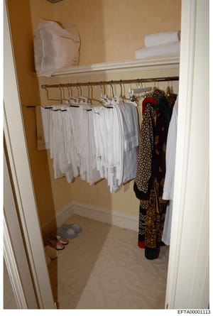 This photograph depicts a walk-in closet or dressing room containing white linens and towels hanging on a rod, decorative patterned garments on the right side, shoes on the floor, and rolled towels on the upper shelf. The image appears to be evidence