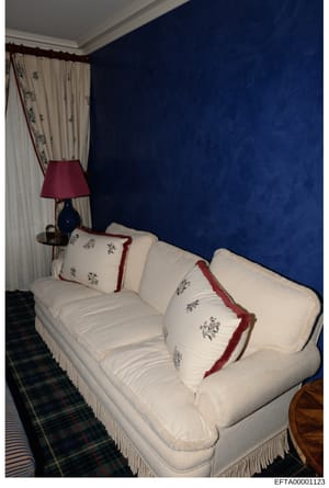 This is an evidence photograph showing the interior of a residential bedroom, likely from one of Epstein's properties. The room features a white upholstered sofa with decorative pillows, navy blue wall paneling, cream and floral patterned curtains wi
