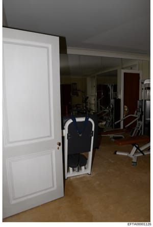 This photograph shows an interior view of a residential fitness/exercise room, featuring various exercise machines including a treadmill, weight machines, and training equipment. The room has dark gray walls, mirrored surfaces, and a wooden floor. Th