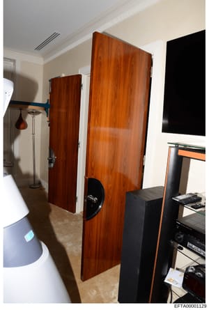 This is a photograph of an interior residential or commercial space showing high-end audio equipment, including two large wooden speaker cabinets and various electronic components. The room features cream-colored walls, recessed lighting, and appears