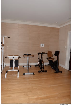 This is a photograph of an indoor home gym or fitness room containing multiple pieces of exercise equipment arranged along a wall. The room features wooden flooring, beige textured wallpaper, and contains at least four different exercise machines inc