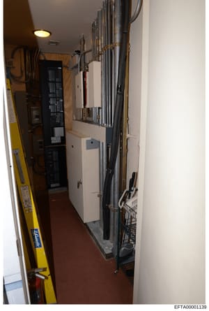 This is an evidence photograph showing an interior electrical/mechanical room containing a main electrical panel, conduit systems, and related building infrastructure. The image documents the physical layout of critical building systems, likely taken