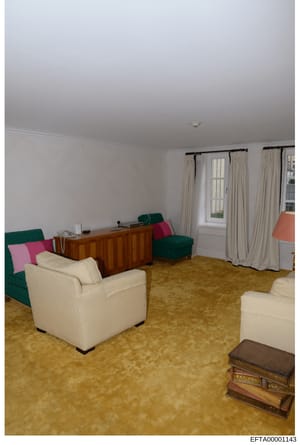 Photograph showing the interior of a living room with cream-colored walls, beige carpet, and mixed furnishings including cream upholstered chairs, green seating, a wooden desk, and cream curtains. The room appears to be part of a larger residential p