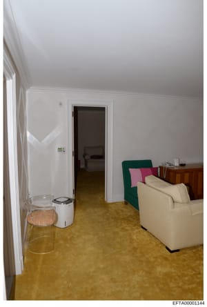 This is a photograph of an interior room, likely a bedroom or sitting area, showing white walls, cork or tile flooring, a cream-colored sofa with green and pink throw pillows, a wooden dresser, and a doorway leading to an adjacent room. The image app