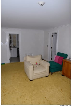 This is a photograph of an interior living space showing a cream-colored armchair, green upholstered chair with pink pillows, wooden dresser, bathroom vanity visible through open doorway, and beige carpeting. The room appears to be part of a residenc
