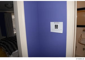 This is an evidence photograph from an Epstein property showing an interior room with distinctive blue walls. A white evidence marker labeled 'BB' is attached to the wall, indicating this photo was taken as part of the official investigation document