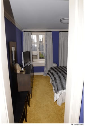 This is a photograph of a modern residential bedroom, likely in a high-rise building based on the elevated city views visible through the windows. The room features contemporary furnishings and décor, including a bed with striped bedding, a desk with