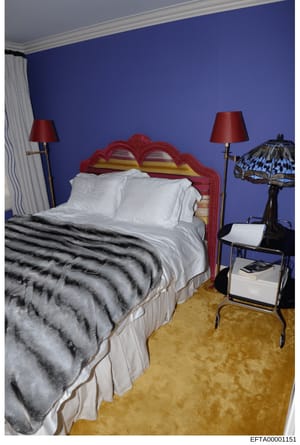 This is a photograph of an upscale bedroom interior featuring a distinctive red ornamental headboard, white bedding with a striped fur throw blanket, and coordinated furnishings. The room has bold blue walls with white crown molding and includes deco