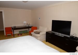 This is an evidence photograph showing the interior of a bedroom, likely from one of Jeffrey Epstein's properties. The room contains a bed with white linens, a desk with red chairs, a cream-colored armchair, a flat-screen television, and various furn