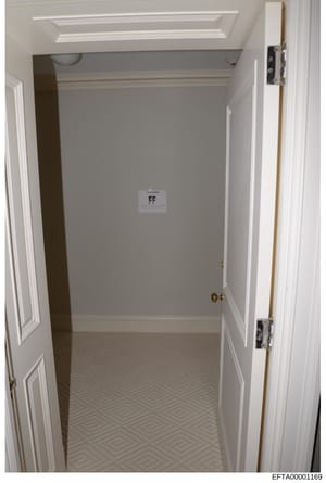 This is a photograph of a narrow, white interior hallway or closet space with minimal furnishings. The room features white-painted walls, recessed ceiling lighting, white trim and baseboards, a patterned floor with a geometric diamond design, and a s