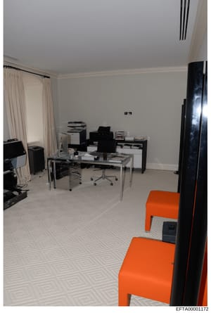 This is a photograph of a modern office interior, showing a workspace with a glass desk, computer equipment, filing systems, and office furniture. The room features white walls, geometric patterned flooring, and includes two distinctive orange chairs